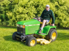 Dennis, smiling, on a green John Deere 255 ride-on lawnmower in front of a tall hedge